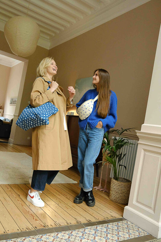 Deux femmes qui discutent, l'une porte le grand sac cabas en denim imprimé cœur, l'autre porte la banane en coton matelassé imprimé little flower, petites fleurs lilas sur fond beige. Accessoires et sacs pour femme en coton.