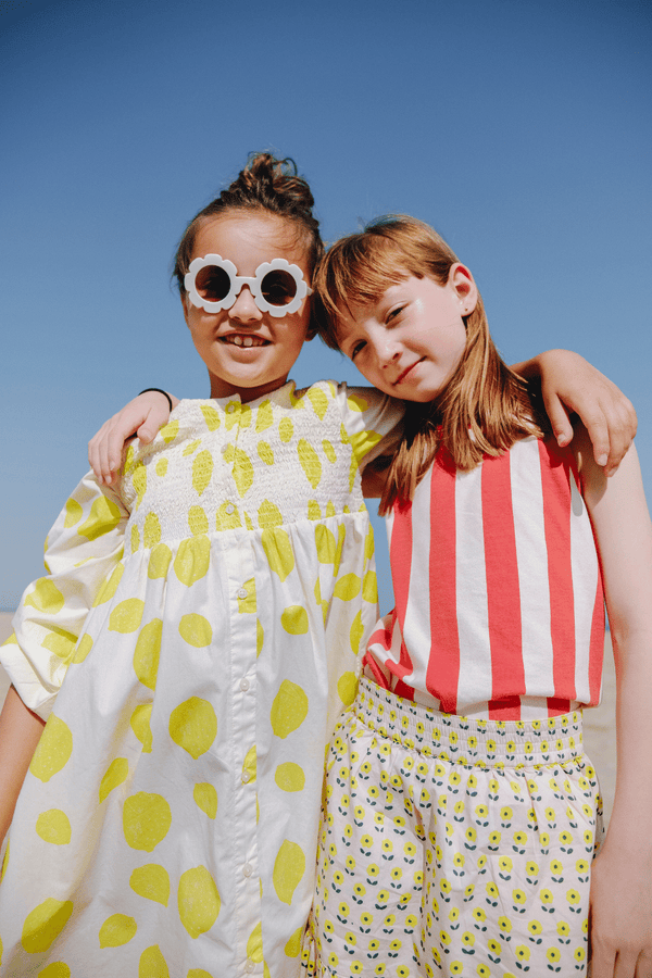 Deux filles jouant sur la plage, l'une portant la robe EDNA motif citron et l'autre le débardeur MARCEL à rayures rouges et la jupe IRMA imprimé petites fleurs jaunes, vêtements en coton bio certifié GOTS style vintage mixte unisexe