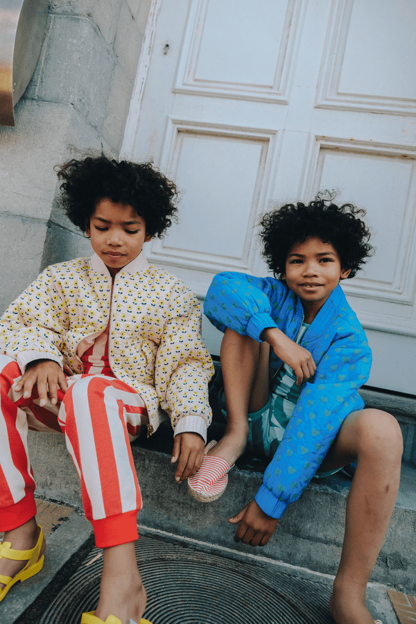 Deux enfants s'amusent, l'un porte un bomber gaby bleu motif cœur vert avec le débardeur marcel et le short kim à rayures vertes, l'autre porte le bomber gaby imprimé petites fleurs jaunes, vêtements en coton bio style vintage