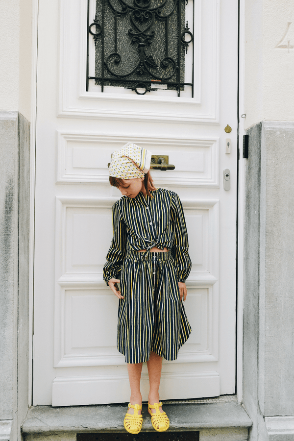 Fille habillé avec l'ensemble chemise SASHA à manches longues et la  jupe LISE coupée au genou avec poches motif rayures bleu marine, rose et jaune, et le foulard BRIGITTE imprimé petites fleurs jaunes, vêtement en coton bio style vintage