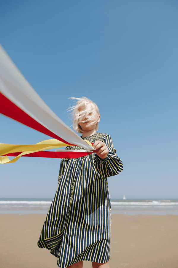 Fille jouant à la plage portant la robe coupe longue à manches longues ALMa à rayures bleu marine rose et jaune vêtements en coton bio certifié GOTS style vintage mixte unisexe