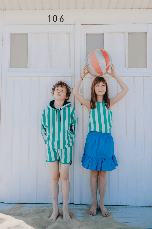 Enfant à la plage, le garçon porte un sweat et le short KIM à rayures vertes, la fille porte le débardeur MARCEL et la jupe IRMA à poches et short intégré couleur bleue et imprimé cœurs verts, vêtements en coton bio certifié GOTS style vintage mixte unisexe