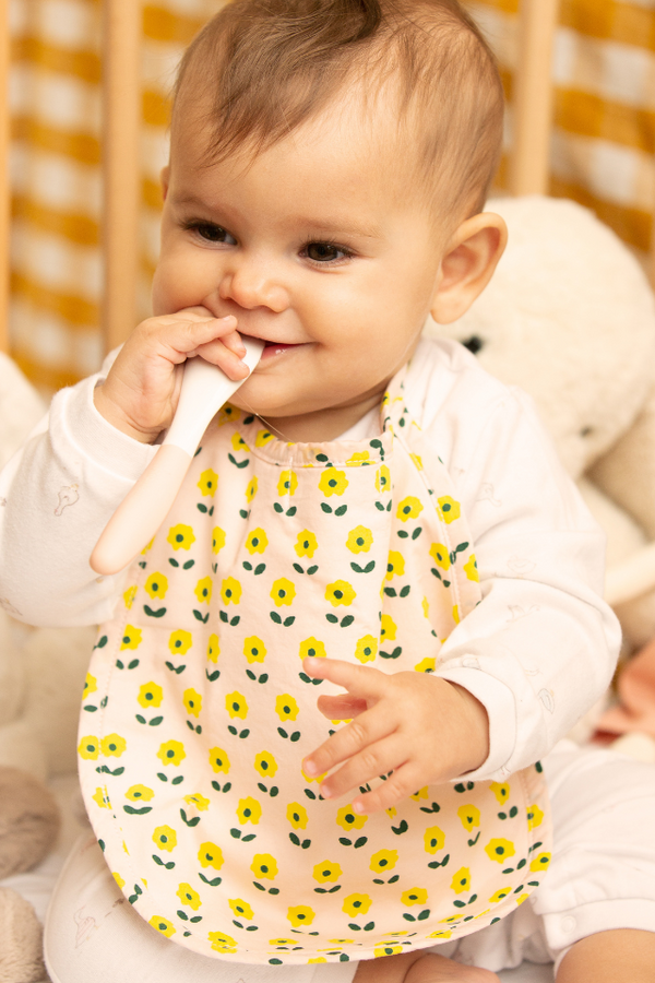 Bébé joyeux jouant avec une petite cuillère, qui porte le bavoir à motif Flower en coton bio de la capsule bébé naissance Baby Care